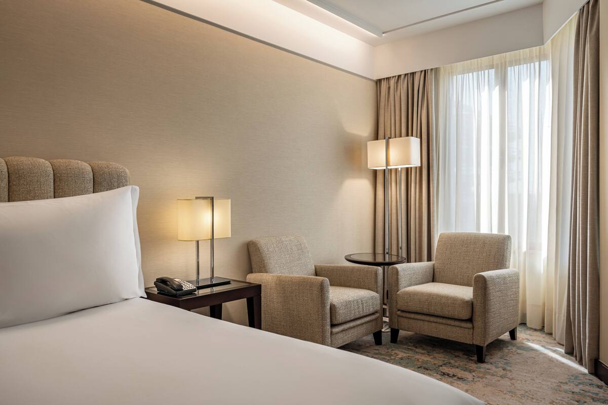 A hotel room with a large bed, two armchairs, a small table with a lamp, a telephone, and a window with curtains.