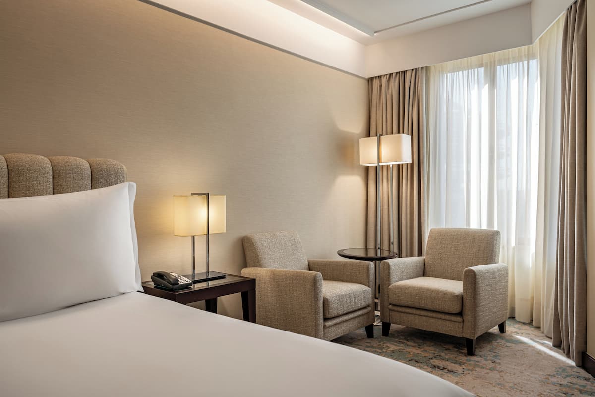 A hotel room with a large bed, two armchairs, a small table with a lamp, a telephone, and a window with curtains.