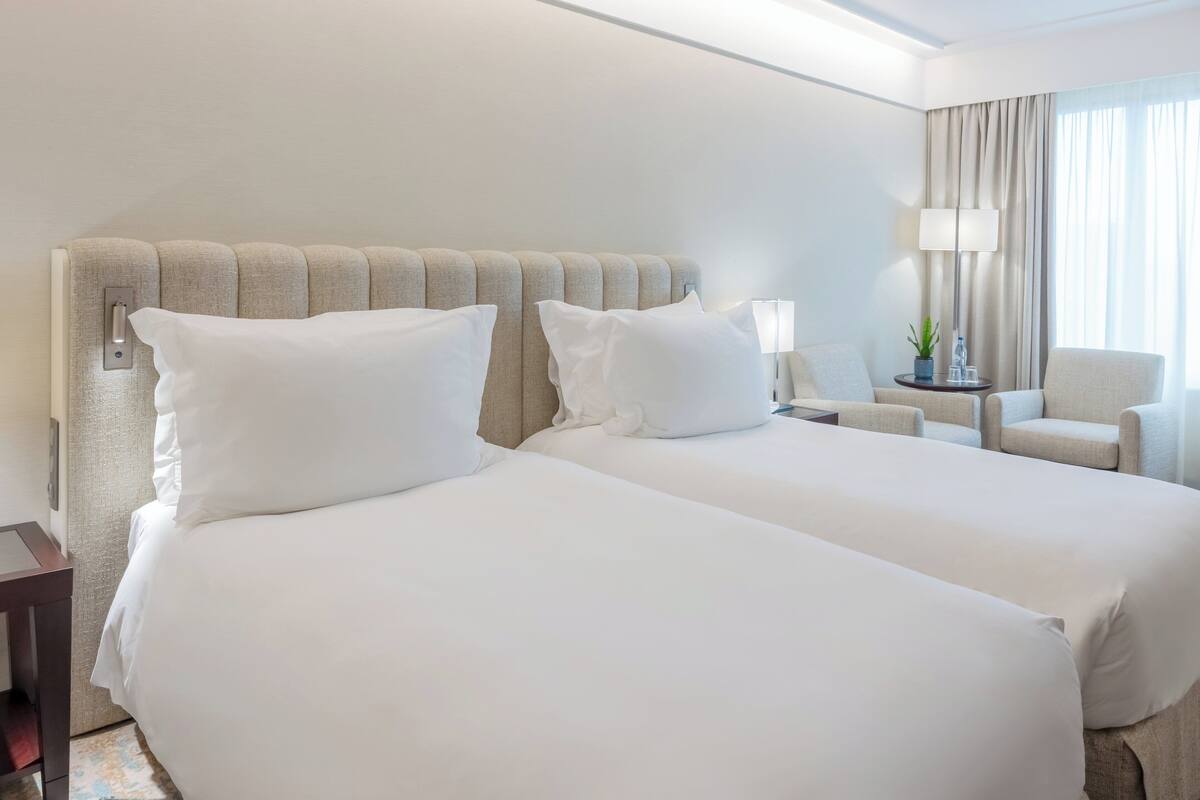 Habitación de hotel con dos camas, una mesita de noche, una lámpara, una silla y un ventanal con cortinas.