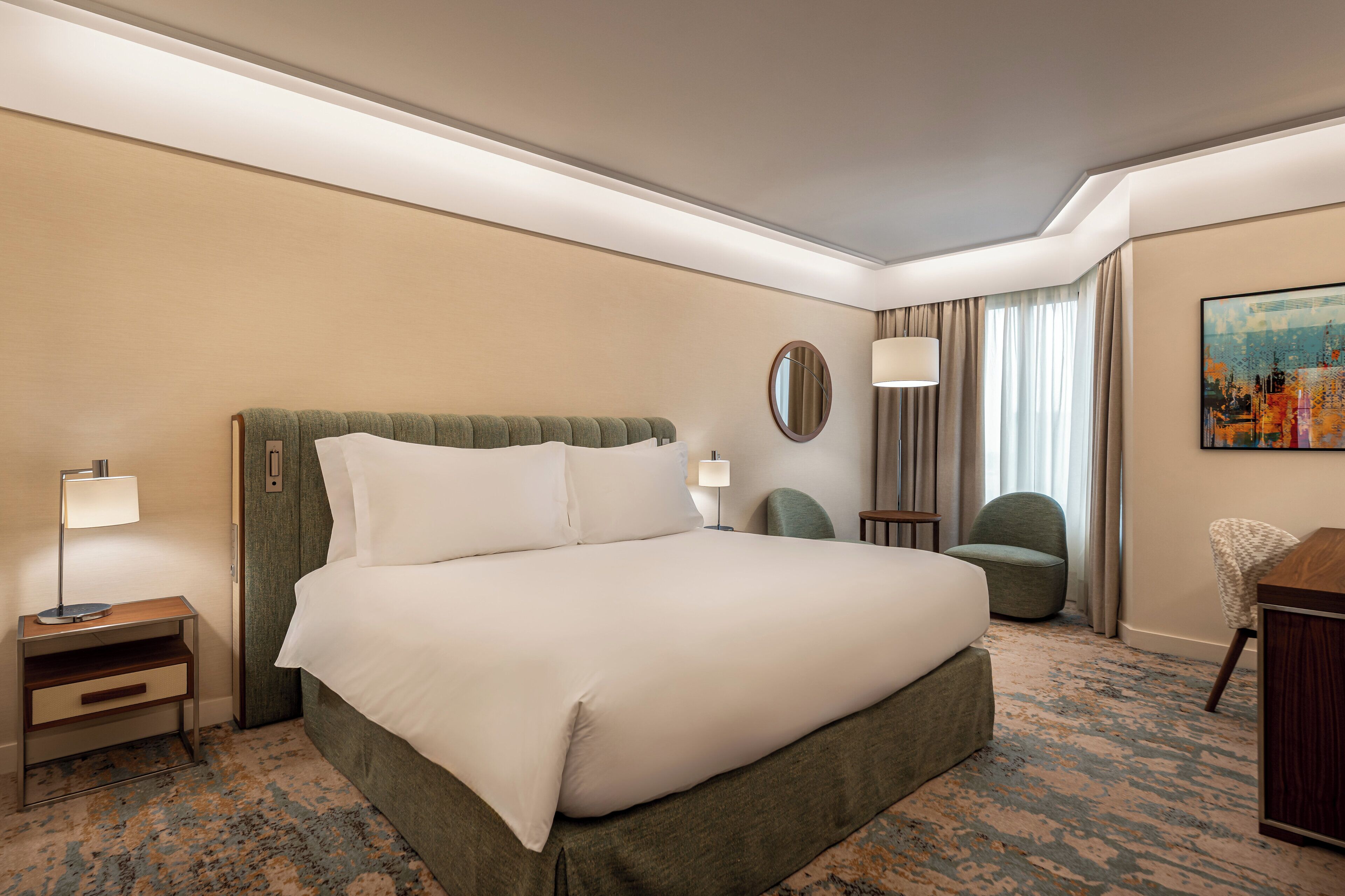 A hotel room with a large bed, two bedside tables, a chair, a desk, and a painting on the wall.