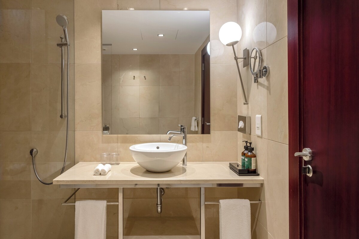 Un baño moderno con un lavamanos color beige, un cabezal de ducha de mano, un espejo con luces y un perchero para toallas.