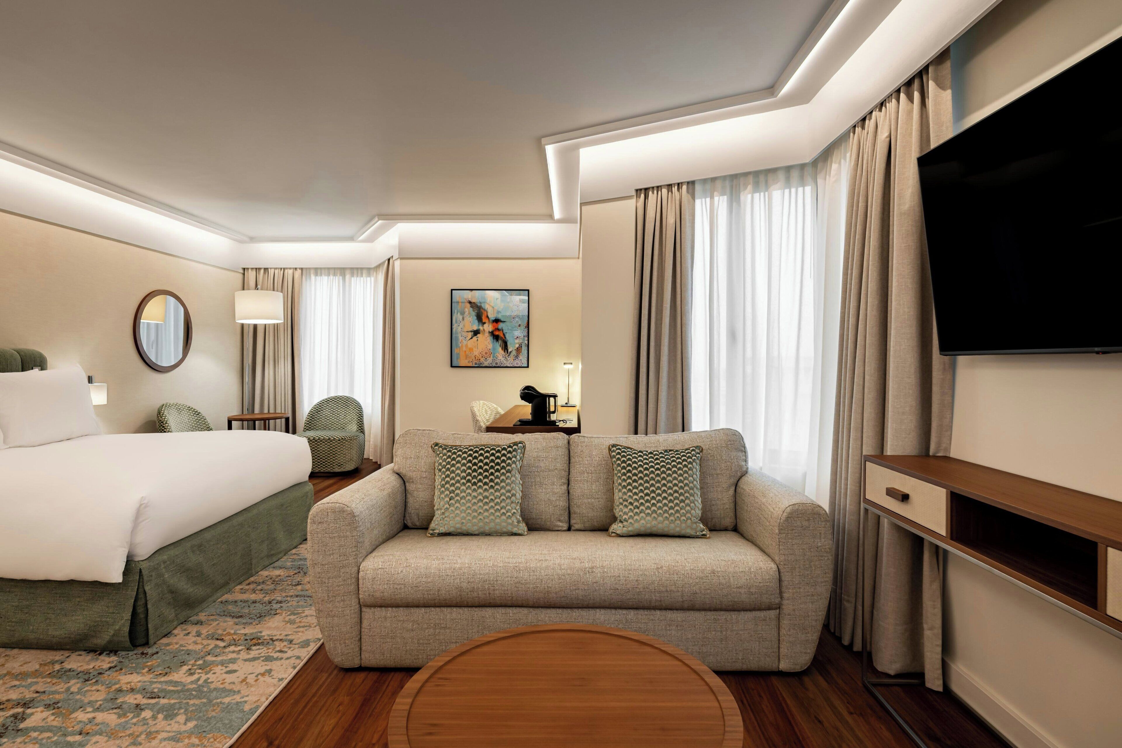 A hotel room with a bed, a sofa, a TV, and a painting on the wall.