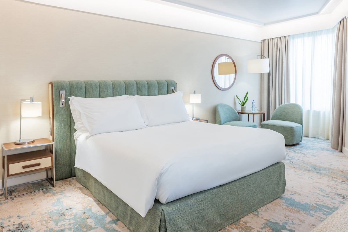 Habitación de hotel con una cama grande, una mesita de noche con lámpara, una silla, una pequeña mesa con una planta y una ventana con cortinas.