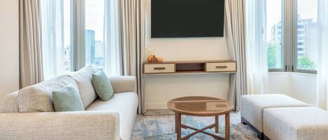 Una habitación de hotel con sofá, una mesita, televisor y ventana con cortinas.