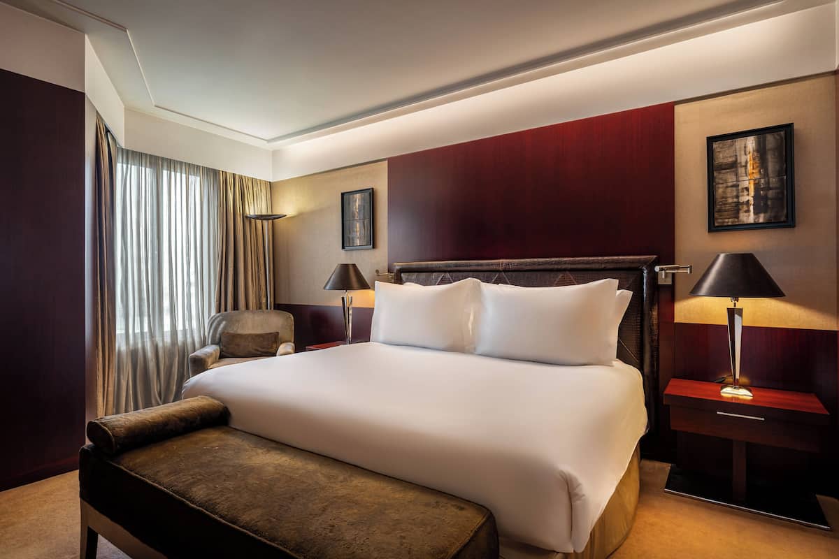 Una habitación de hotel con una cama grande, una mesita de noche con lámpara, una silla y una ventana con cortinas.