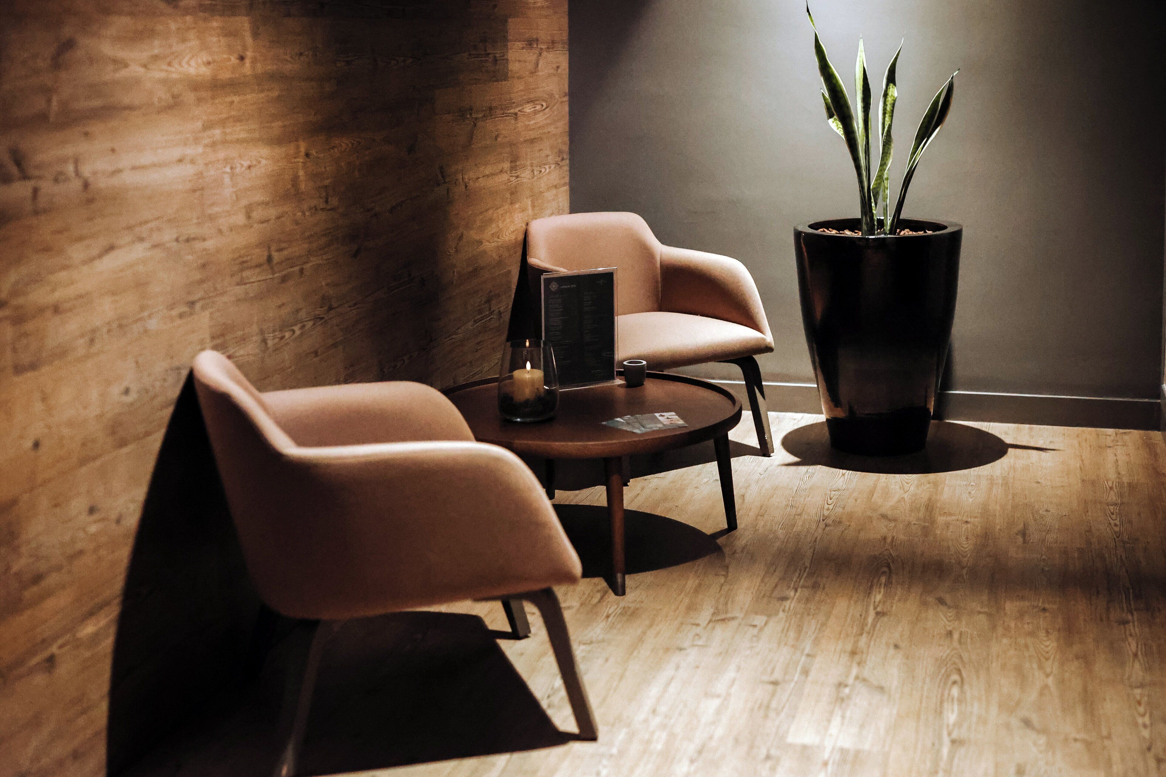 A modern room with a wooden floor, two cushioned chairs, a small round table, a potted plant, and a wall-mounted shelf with items.