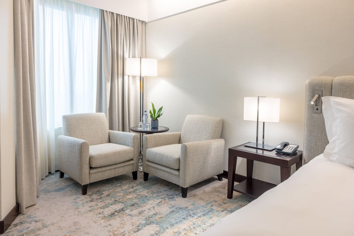 A hotel room with two armchairs, a small table with a lamp, a bedside table with a phone, and a bed.