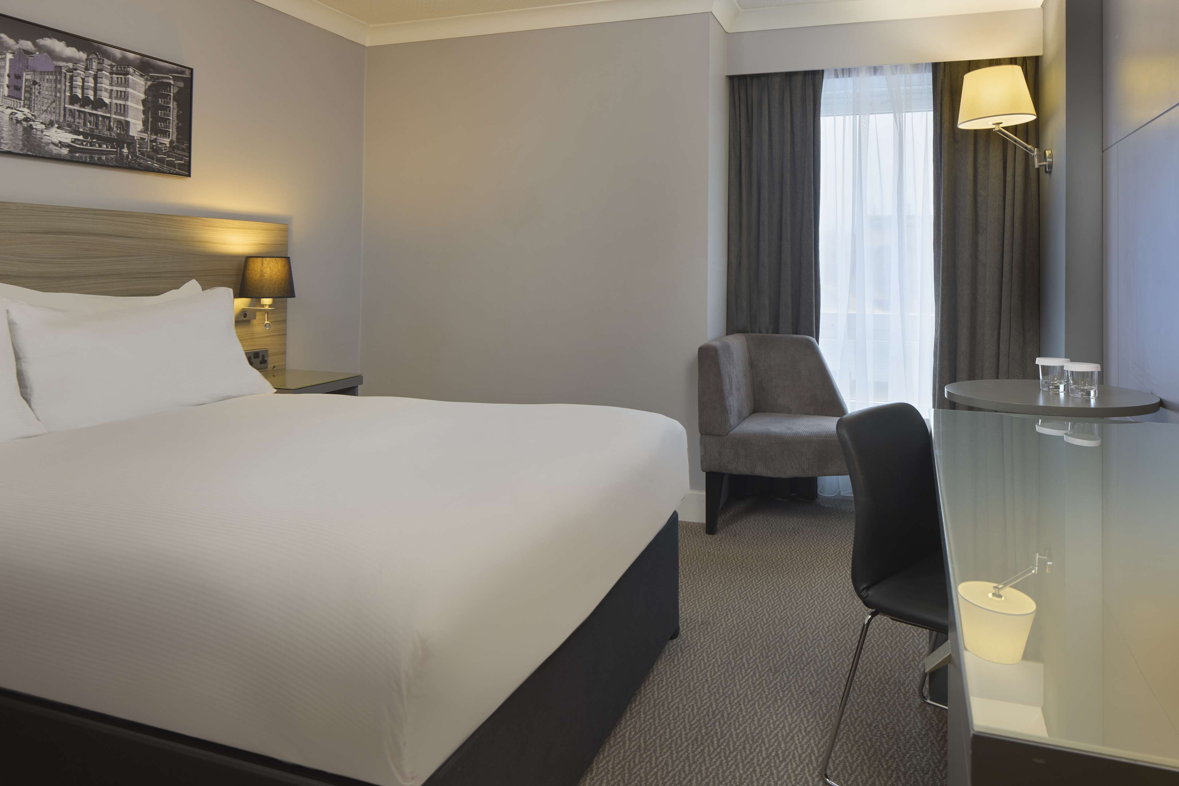 Quarto de hotel com uma cama grande, uma cadeira, uma mesa com abajur e um quadro na parede.