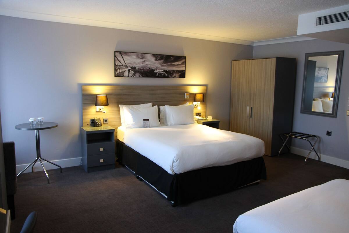 A hotel room with a large bed, bedside tables, a small table, a wardrobe, and a mirror.