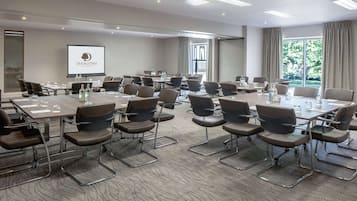 A conference room with multiple tables and chairs, a projector screen displaying a logo, and large windows with curtains.