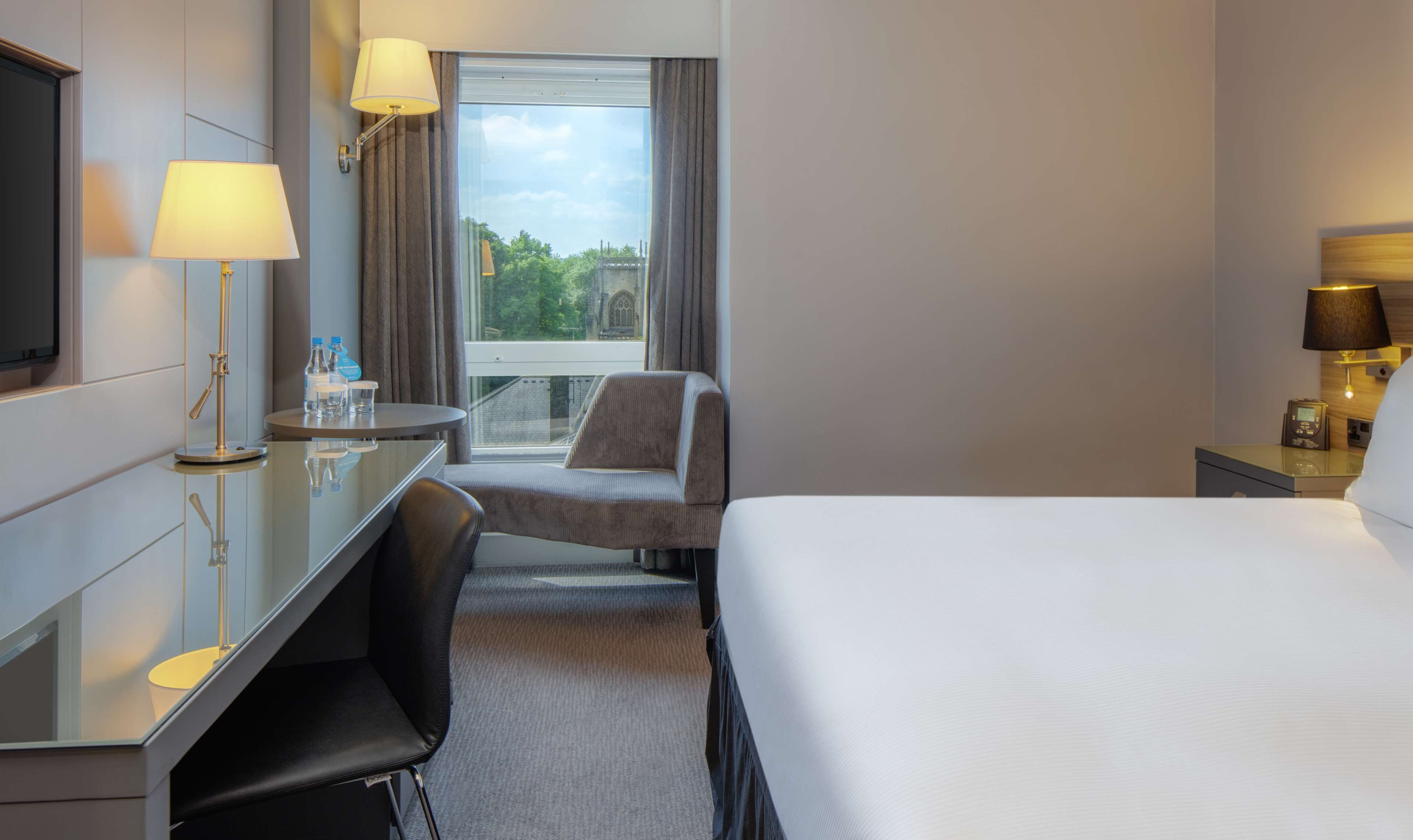 A hotel room with a bed, a desk with a chair, a TV, and a window with curtains.