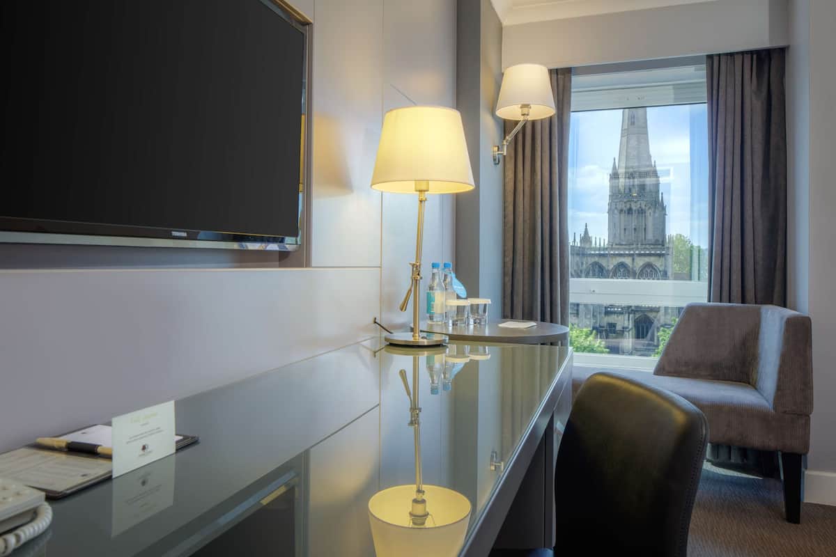 A hotel room with a desk, a chair, a TV, and a window with a view of a cathedral.