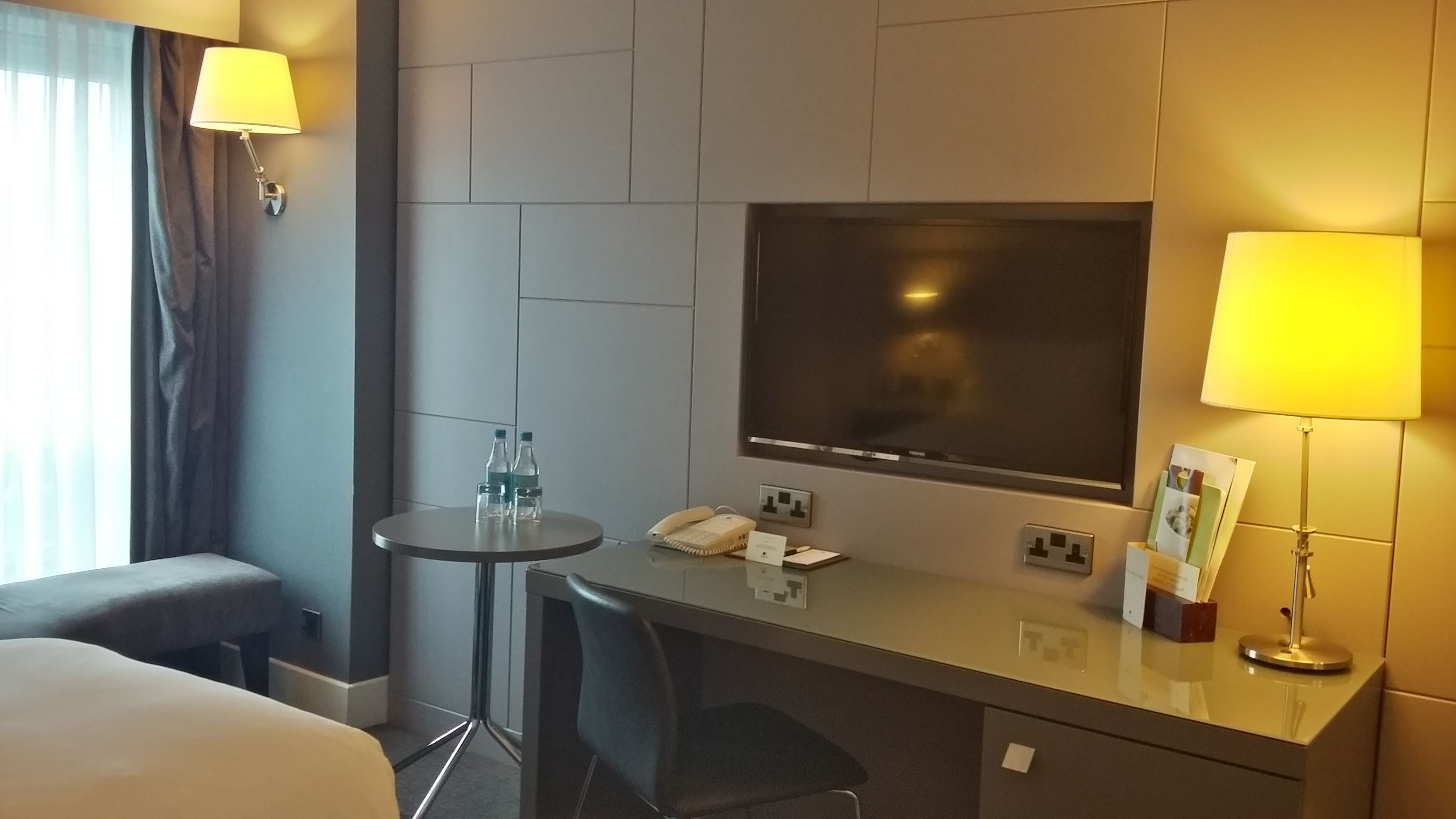 Habitación de hotel con cama, escritorio, silla, televisor, lámpara y ventana con cortinas.