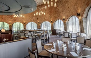 Un restaurante espacioso con techos altos, paredes de ladrillo visto y amplias ventanas en arco que dejan entrar la luz natural. Las mesas están ordenadas con sillas, y el espacio está bien iluminado con lámparas colgantes.