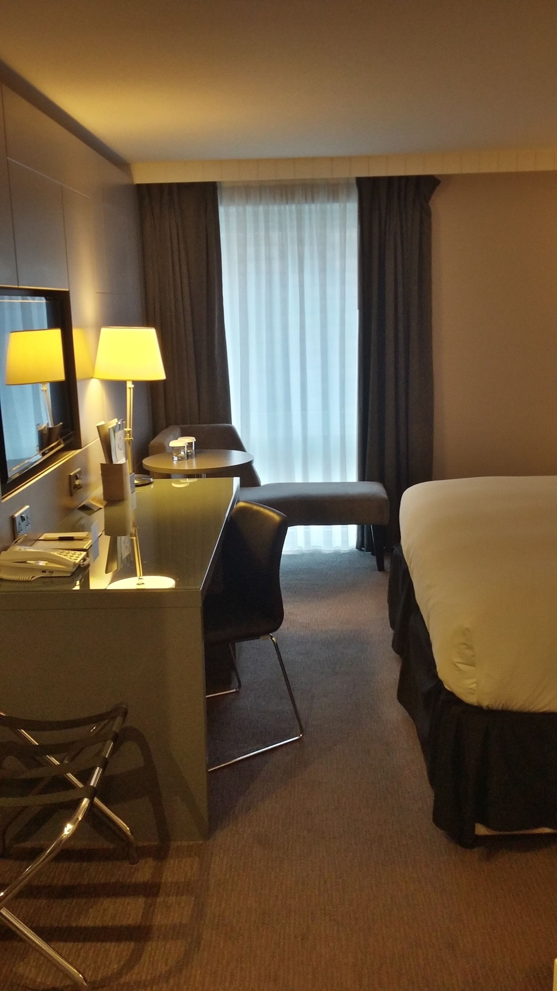 Una habitación de hotel con una cama, un escritorio con dos lámparas, una silla, una mesita y una ventana con cortinas.
