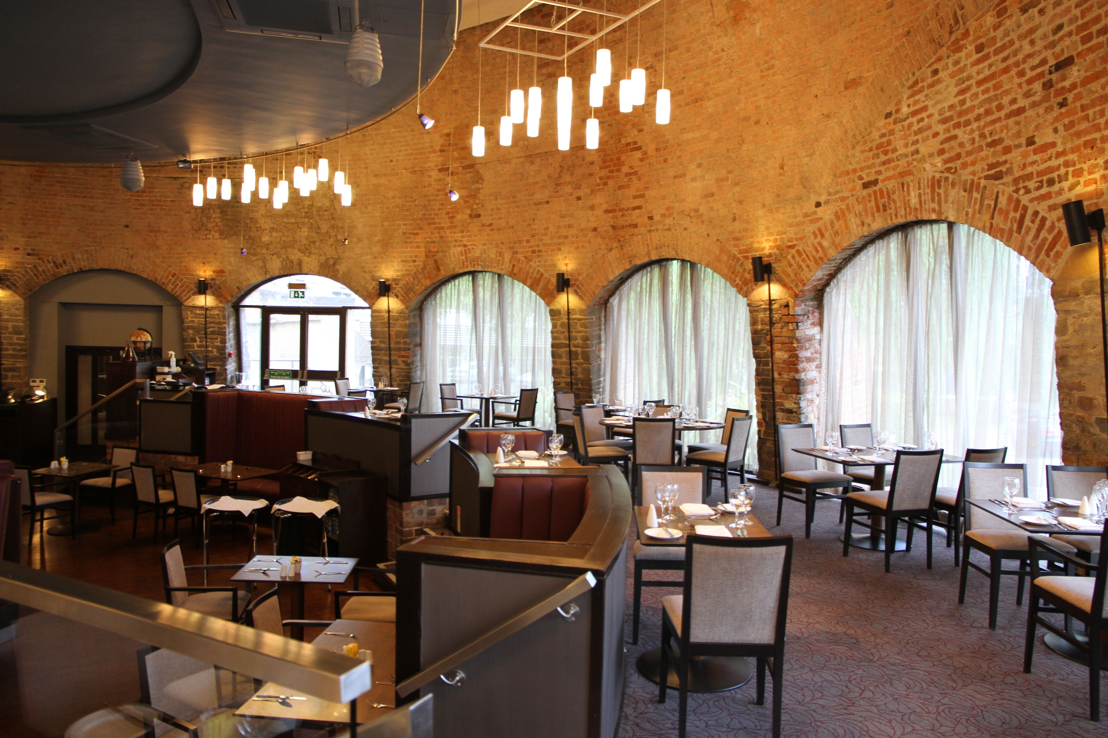 Un restaurante con paredes de ladrillo, ventanas arqueadas e iluminación moderna. Las mesas están preparadas con manteles blancos y cubertería.