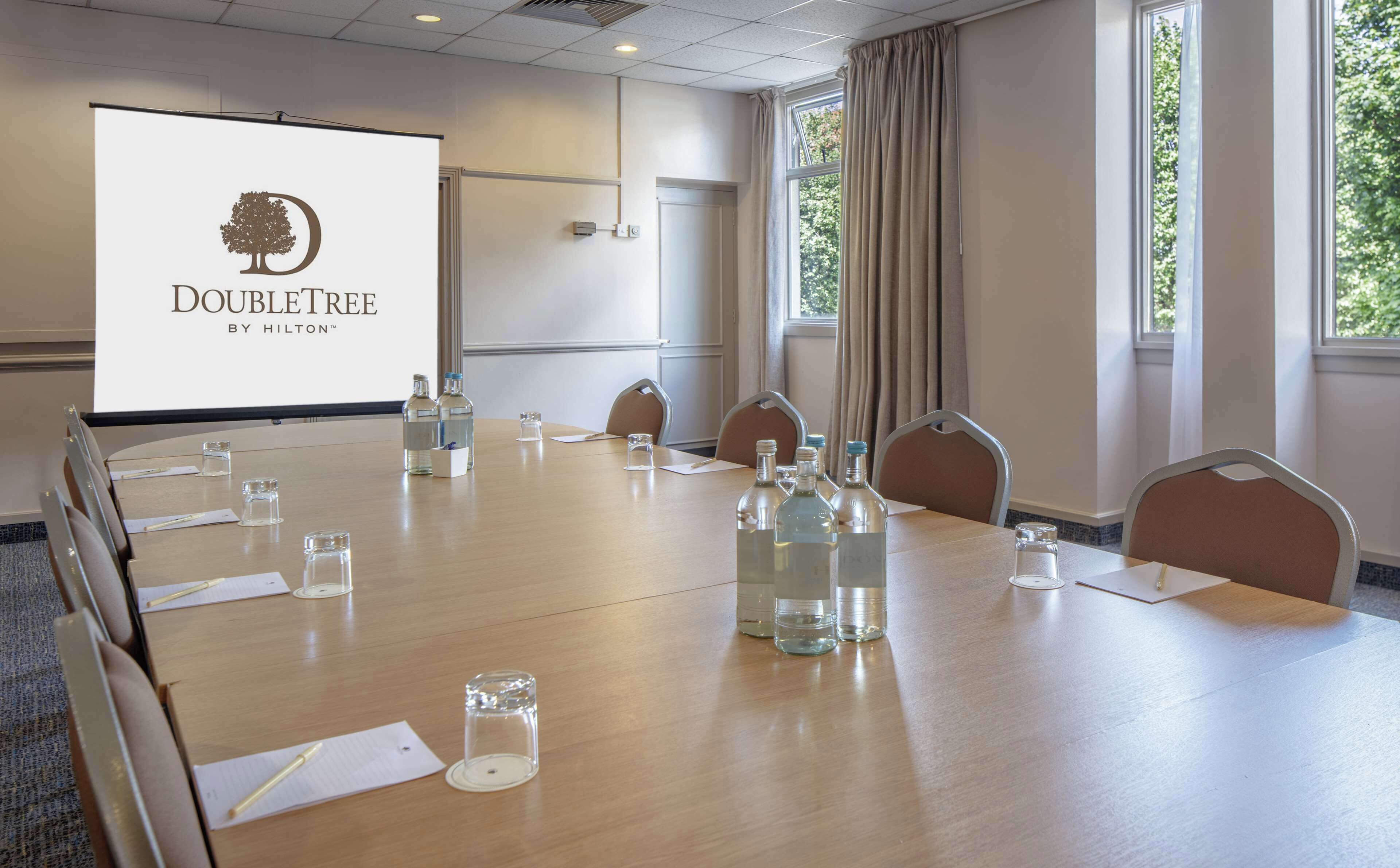 A conference room with a large screen displaying 'DOUBLETREE BY HILTON', a long table with chairs, water bottles, and glasses.