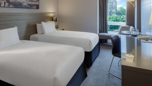 A hotel room with two beds, a desk with a chair, a window with a view of a cathedral, and a painting on the wall. - Doubletree by Hilton Bristol (Bristol)