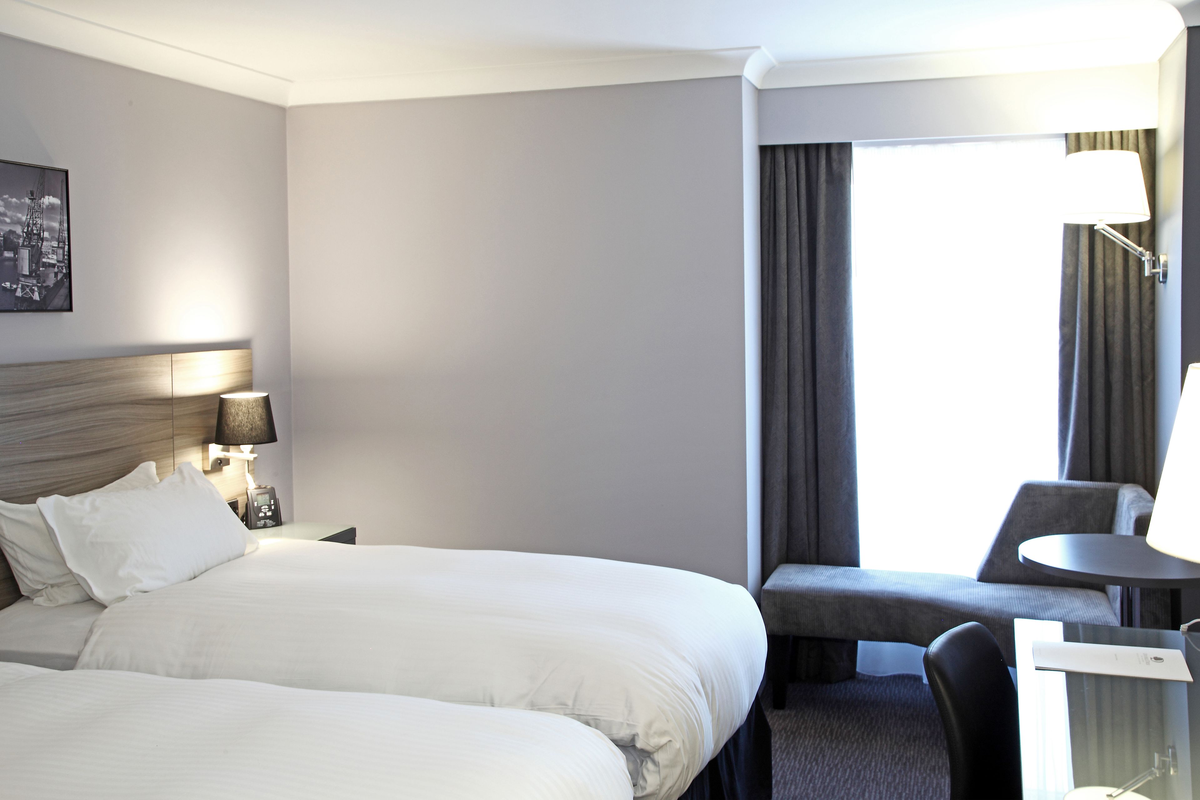 Ein Hotelzimmer mit zwei Betten, einem Schreibtisch, einem Stuhl, einer Lampe und einem fenster mit Vorhängen.