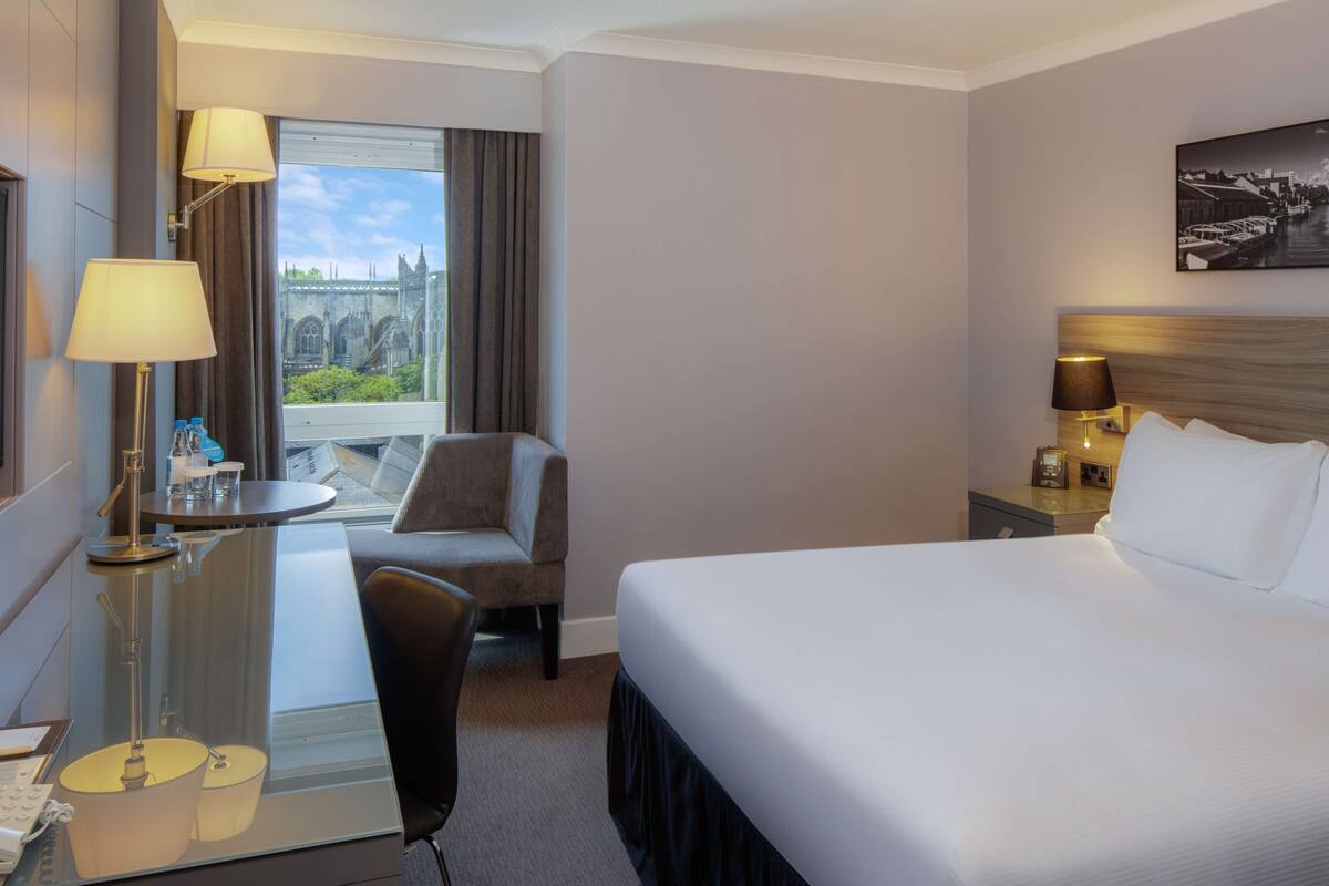 A hotel room with a bed, a desk with a lamp, a chair, a TV, and a window with a view of a bridge.