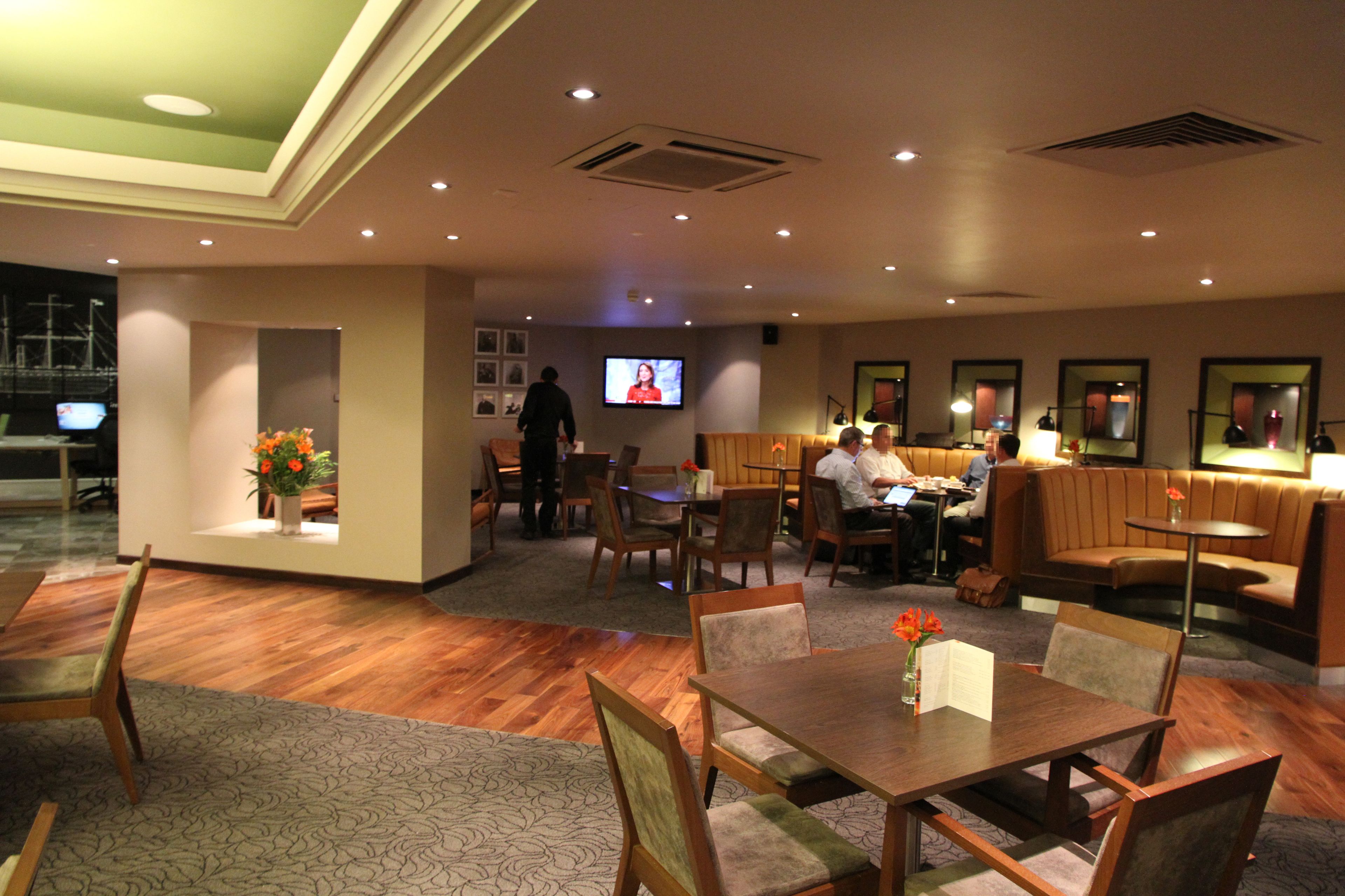 A hotel lobby with wooden flooring, a reception desk, seating areas, and a television screen.
