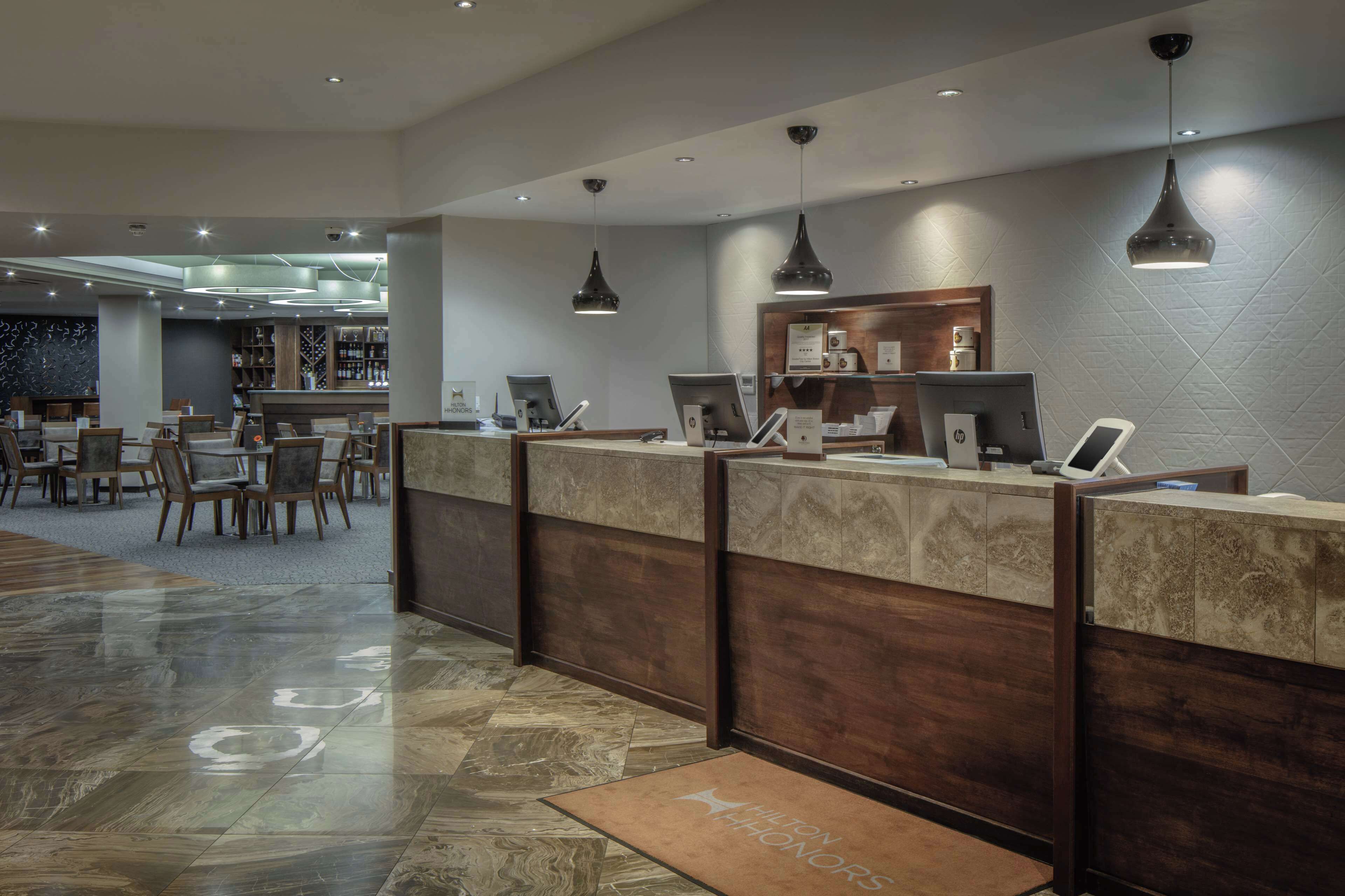 Zona de recepción de un hotel con mostrador, computadoras y un área de estar con mesas y sillas.
