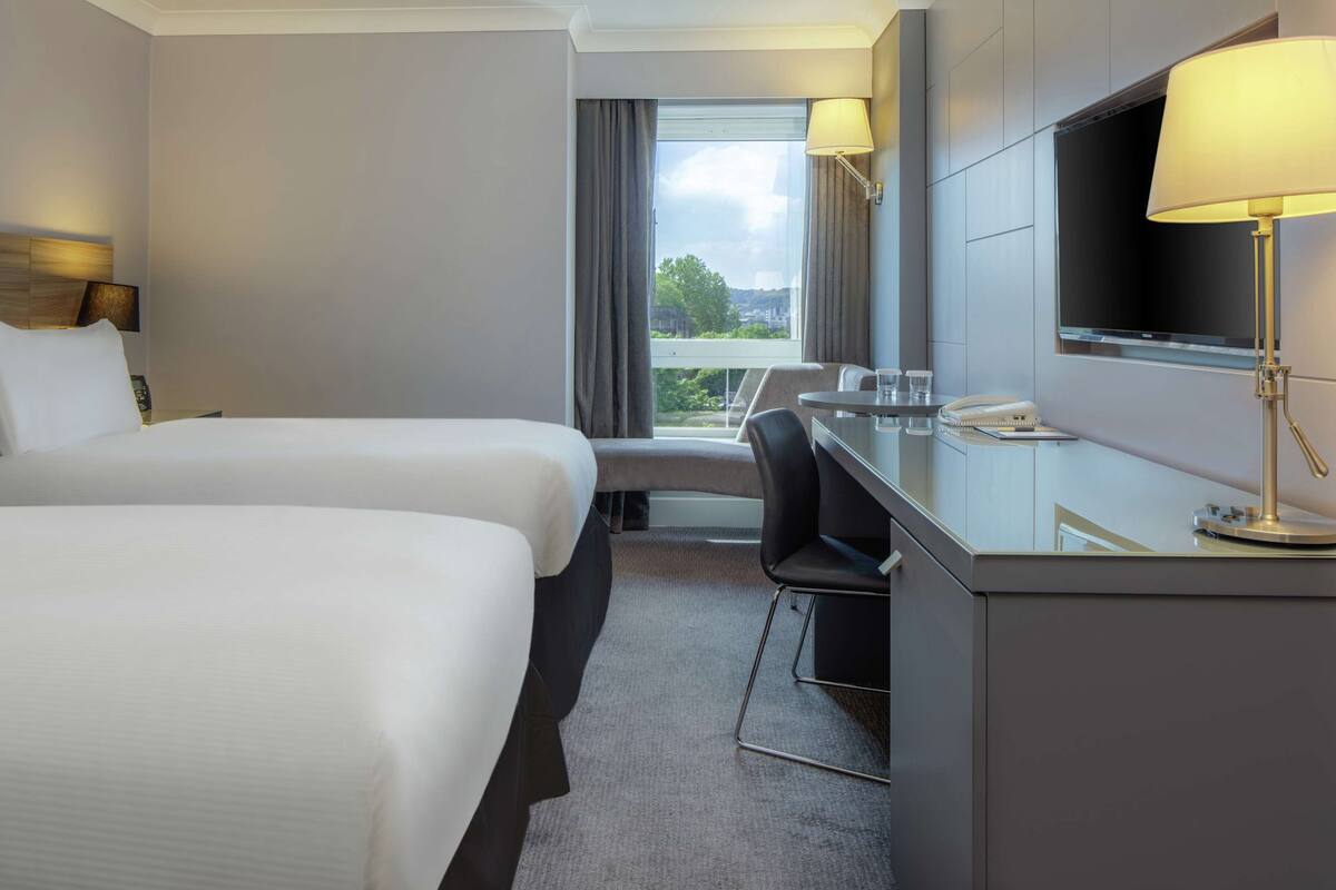 A hotel room with two beds, a desk, a chair, a TV, and a window with a view.