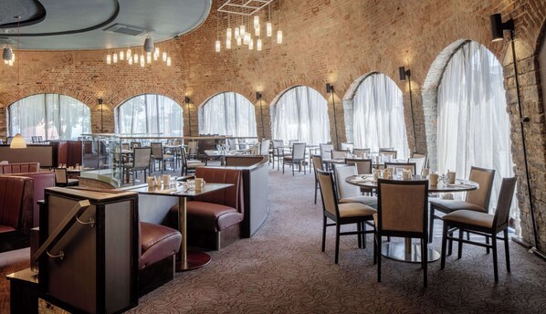 A restaurant with brick walls, large arched windows, and modern lighting. Tables and chairs are arranged for dining.
