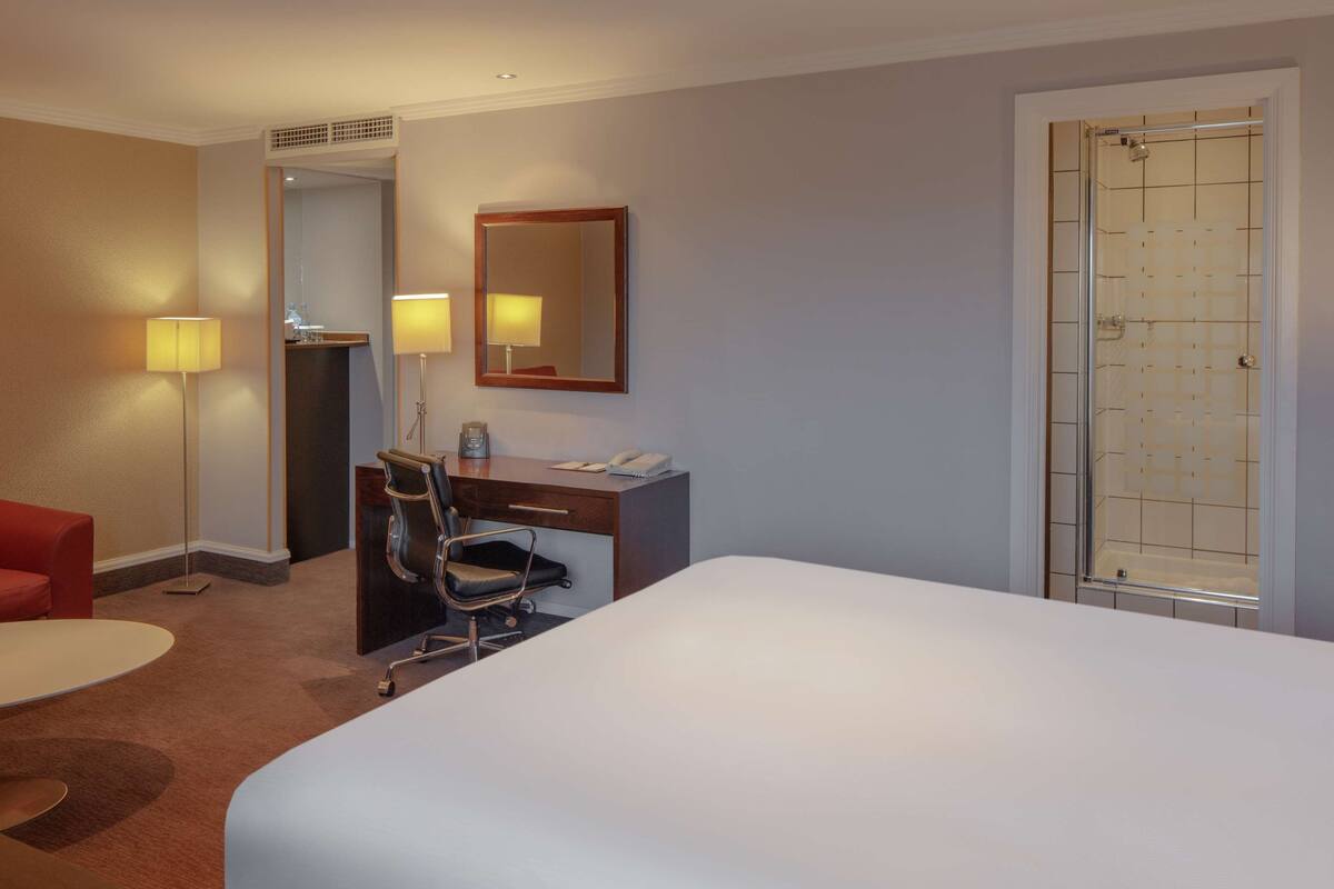A hotel room with a bed, a desk, a chair, a sofa, a lamp, a mirror, and a shower.