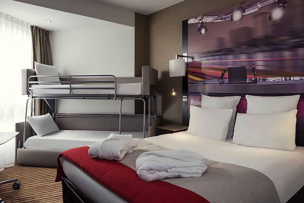 A hotel room with a bunk bed, a large bed with pillows, a headboard with a cityscape mural, and a bedside table with a lamp.