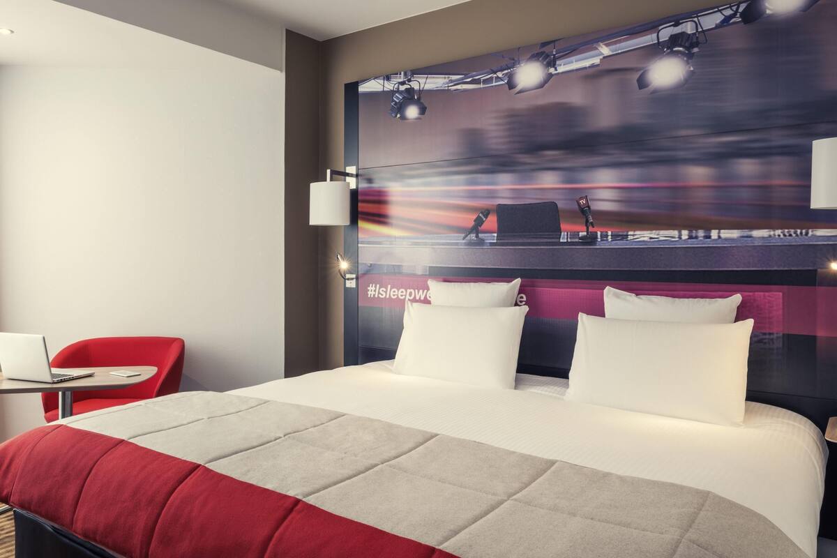 A hotel room with a large bed, two white pillows, a red chair, a desk with a laptop, and a wall with a TV and microphone.