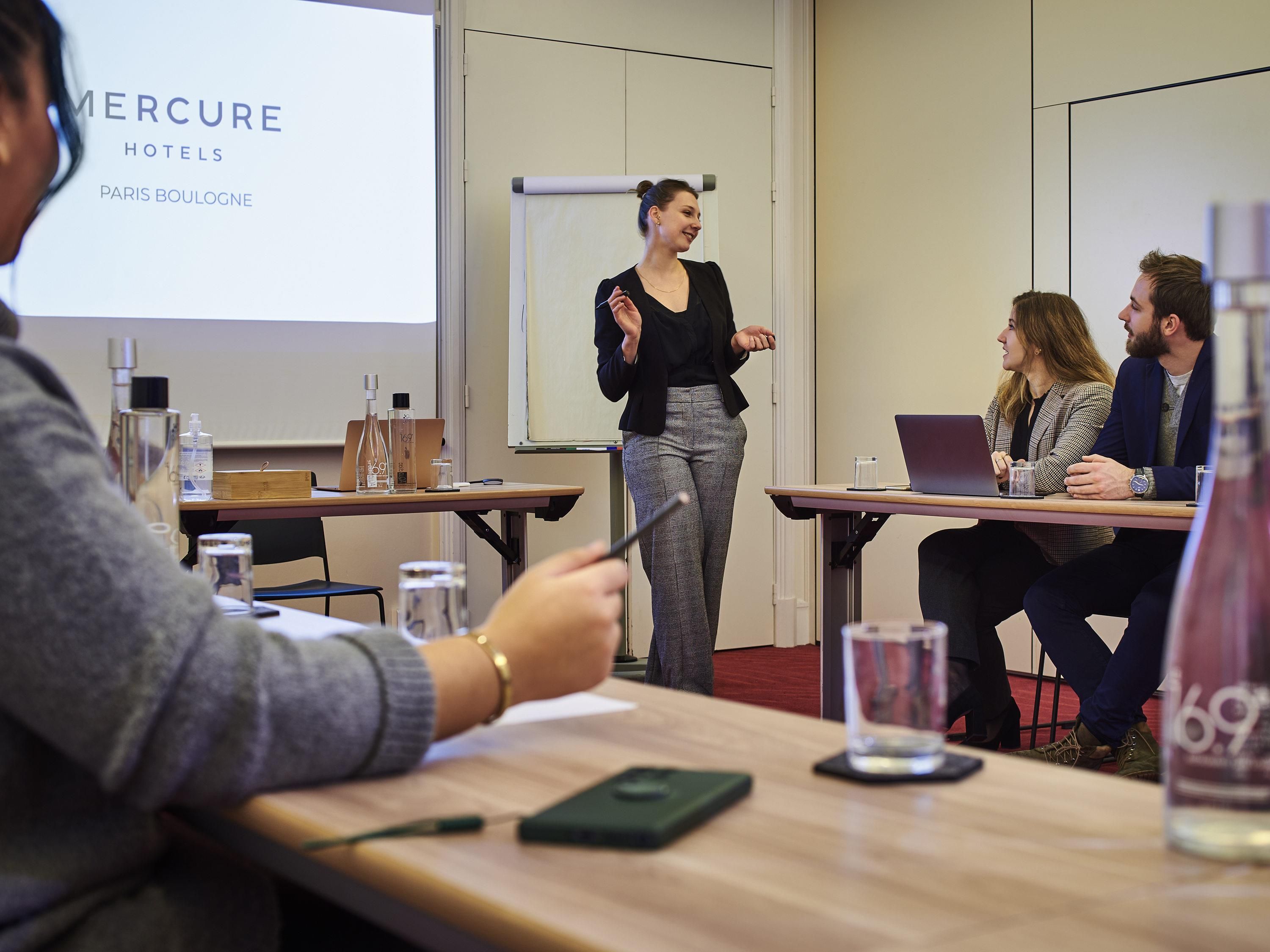 Un salón de conferencias con una pantalla mostrando 'MERCURE HOTELS PARIS BOULOGNE', un presentador y asistentes sentados en mesas con computadoras portátiles.