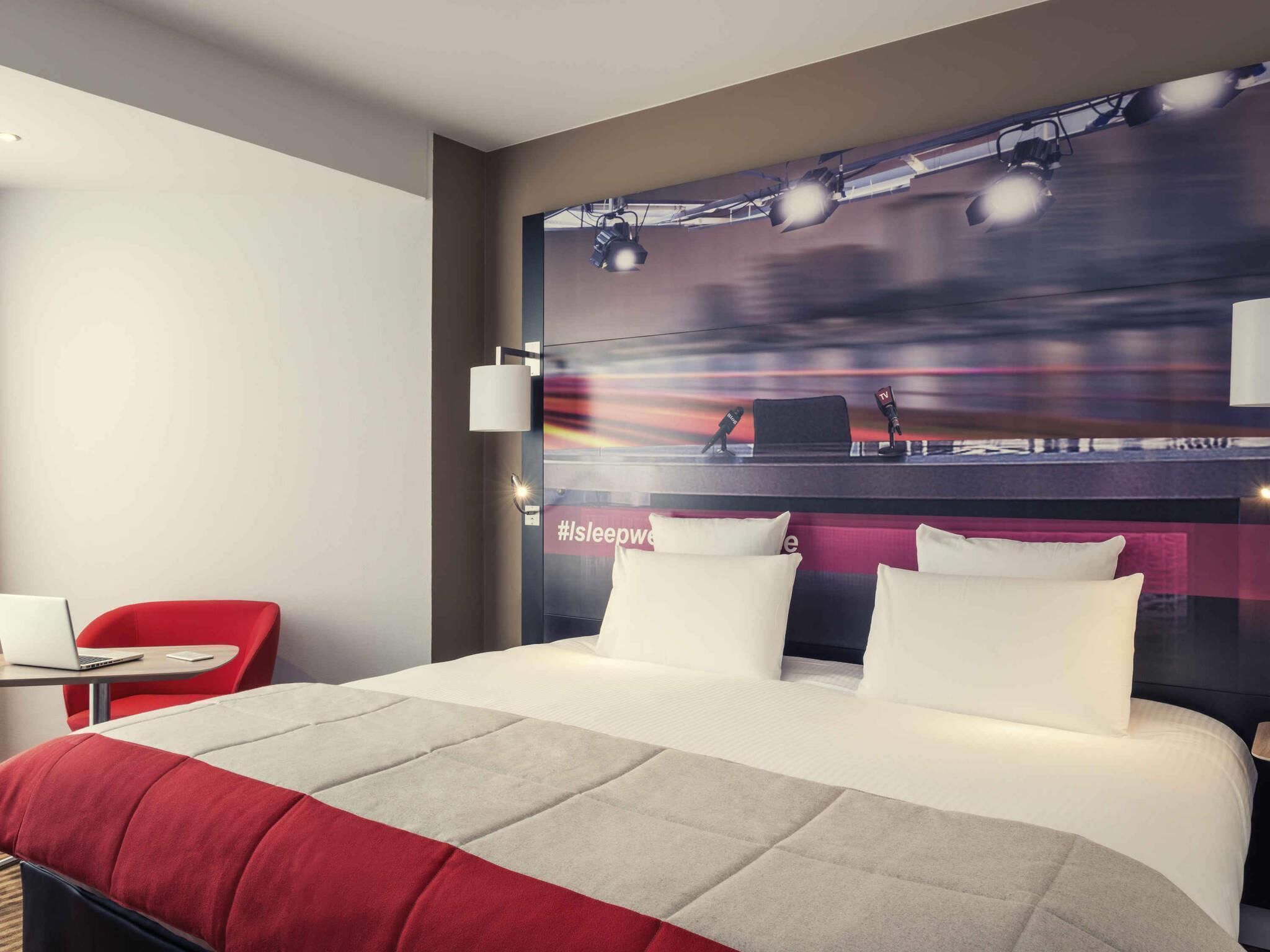 Une chambre d’hôtel avec un grand lit, un bureau avec un ordinateur portable, une chaise rouge et un mur sur lequel sont fixés un téléviseur et un microphone.