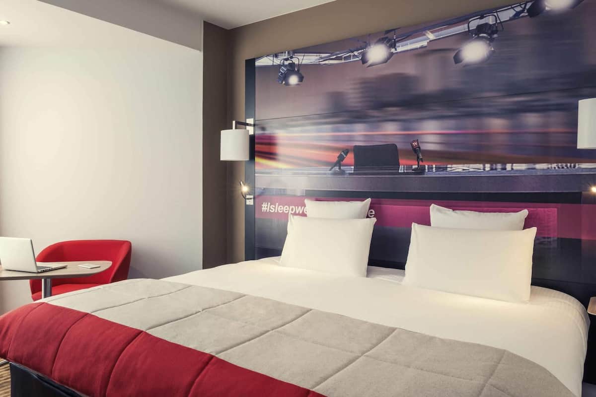 Une chambre d’hôtel avec un grand lit, un bureau avec un ordinateur portable, une chaise rouge et un mur sur lequel sont fixés un téléviseur et un microphone.