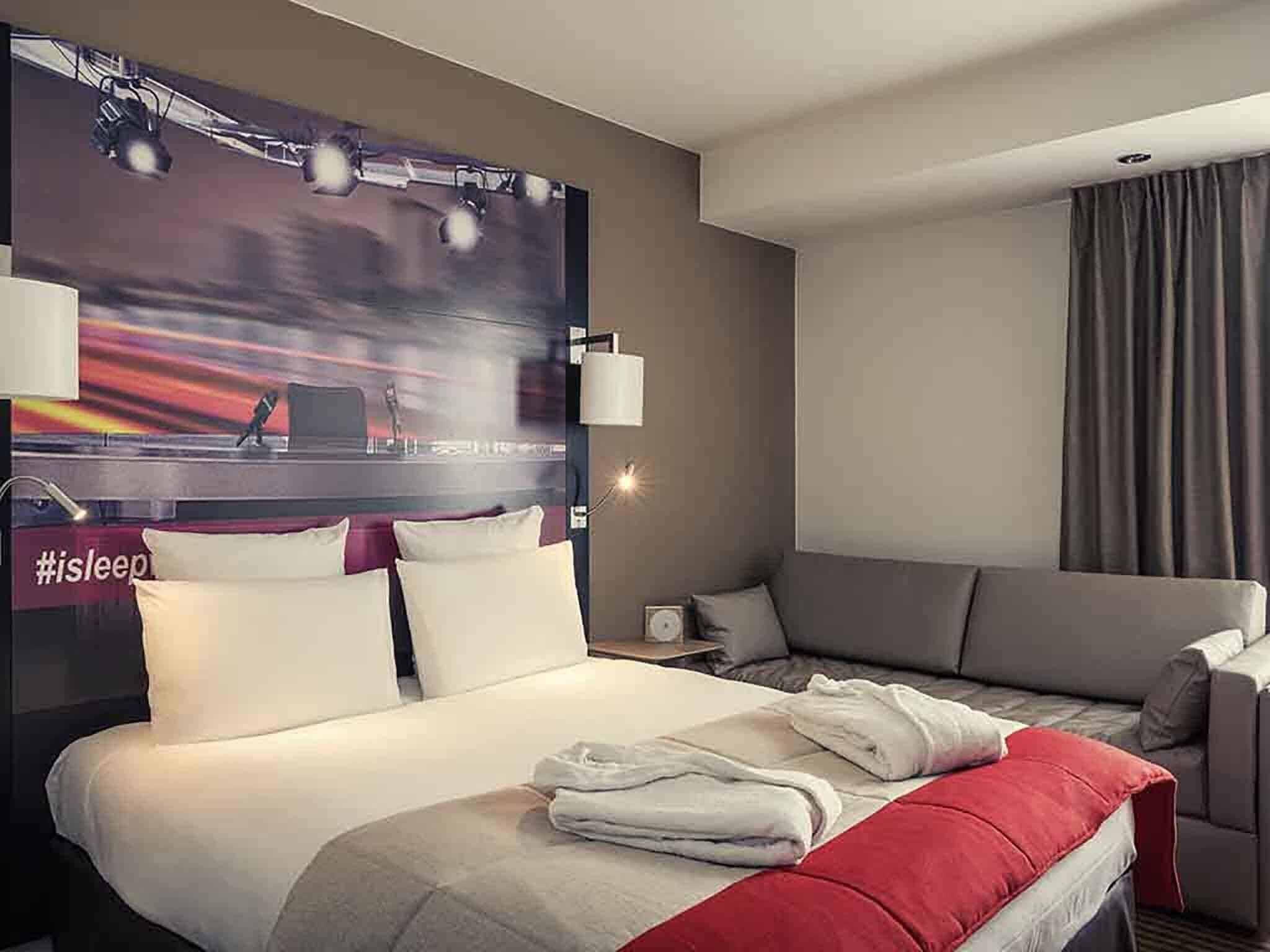 Une chambre d’hôtel avec un lit, un canapé, un téléviseur fixé au mur et une fenêtre avec des rideaux.