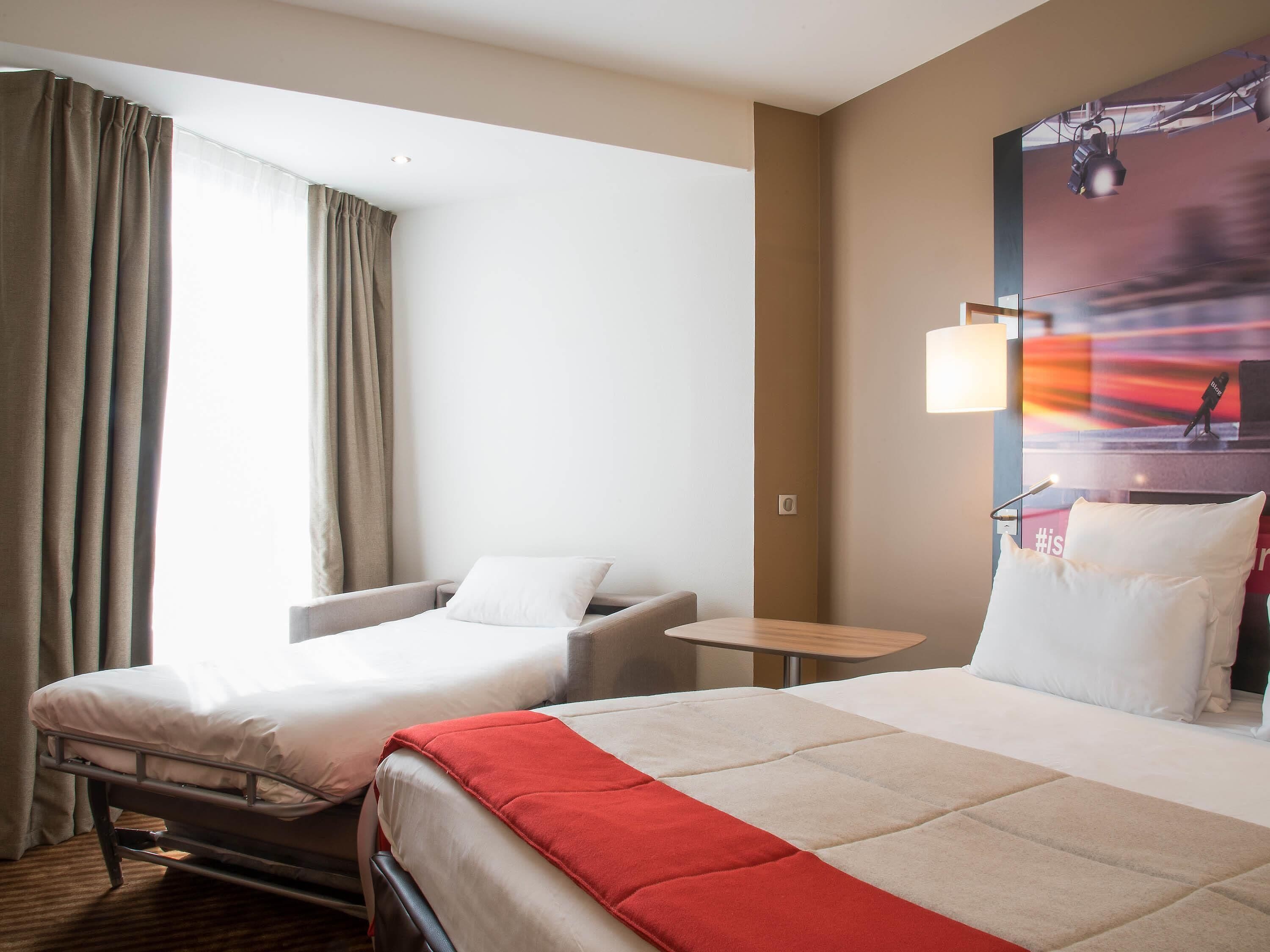 Ein Hotelzimmer mit zwei Betten, einem kleinen Tisch, einem Stuhl und einem großen Fenster mit Vorhängen.