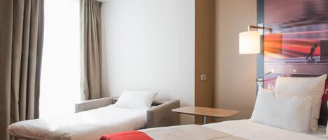 Ein Hotelzimmer mit zwei Betten, einem kleinen Tisch, einem Stuhl und einem großen Fenster mit Vorhängen.