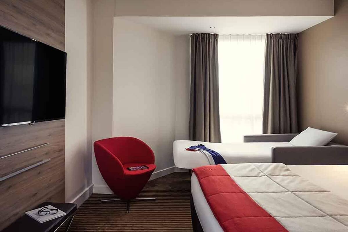 A hotel room with a bed, a red chair, a TV, and a window with curtains.