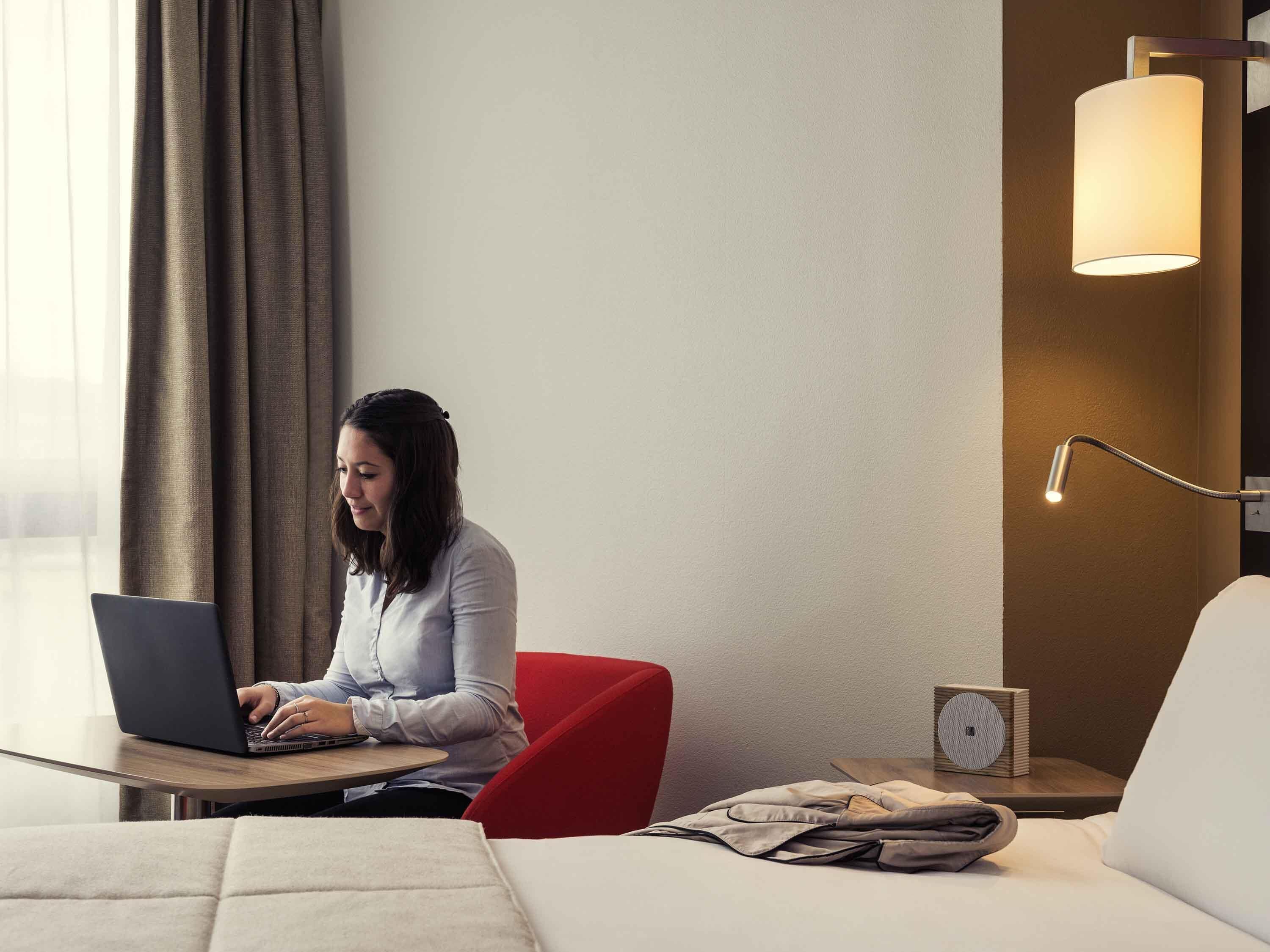 A hotel room with a bed, a desk with a laptop, a red chair, a lamp, and a small speaker.