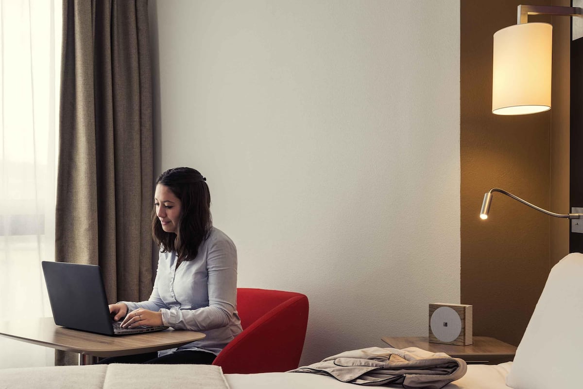 A hotel room with a bed, a desk with a laptop, a red chair, a lamp, and a small speaker.