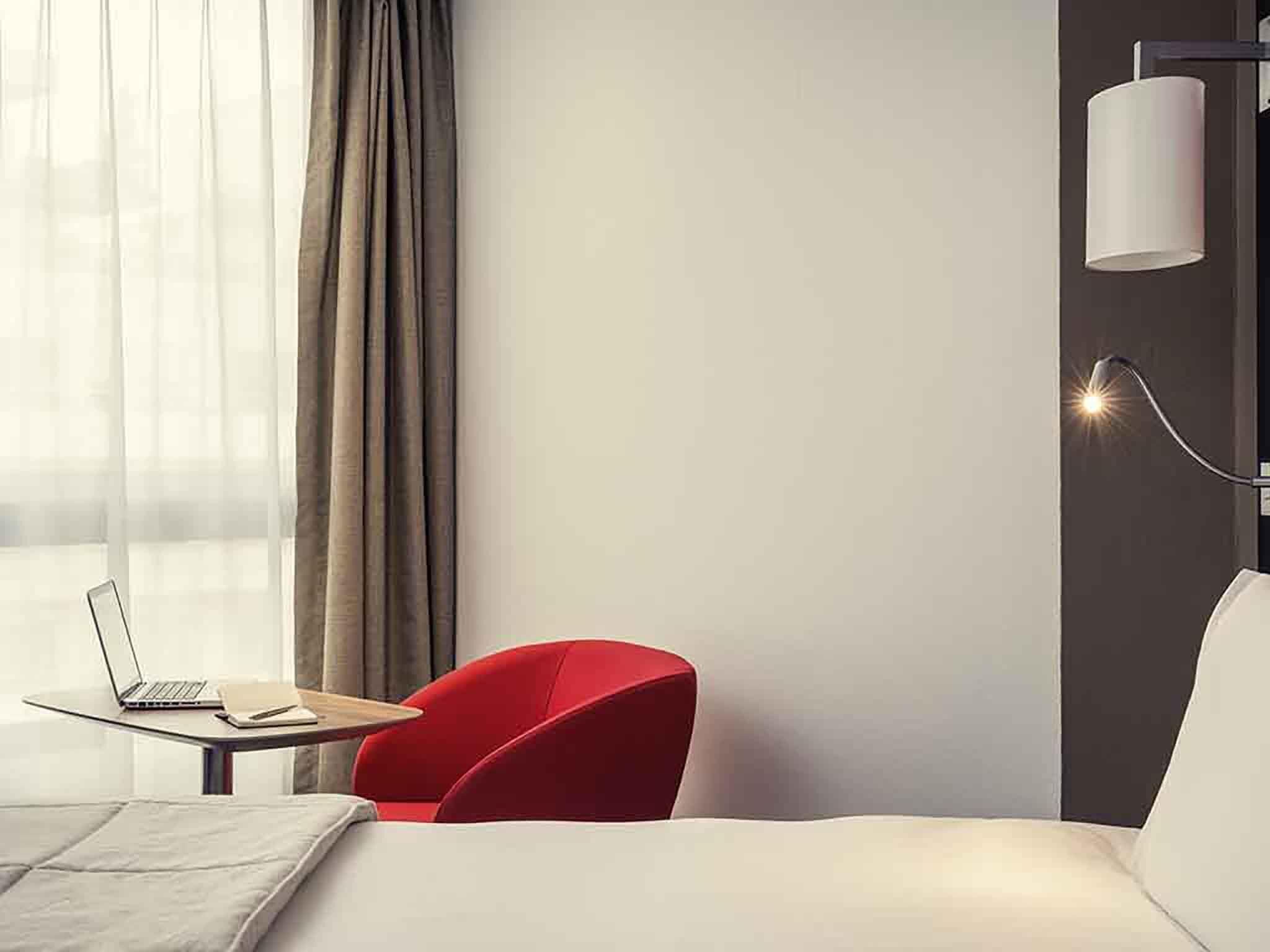 A modern hotel room with a bed, a red armchair, a desk with a laptop, and a wall-mounted lamp.