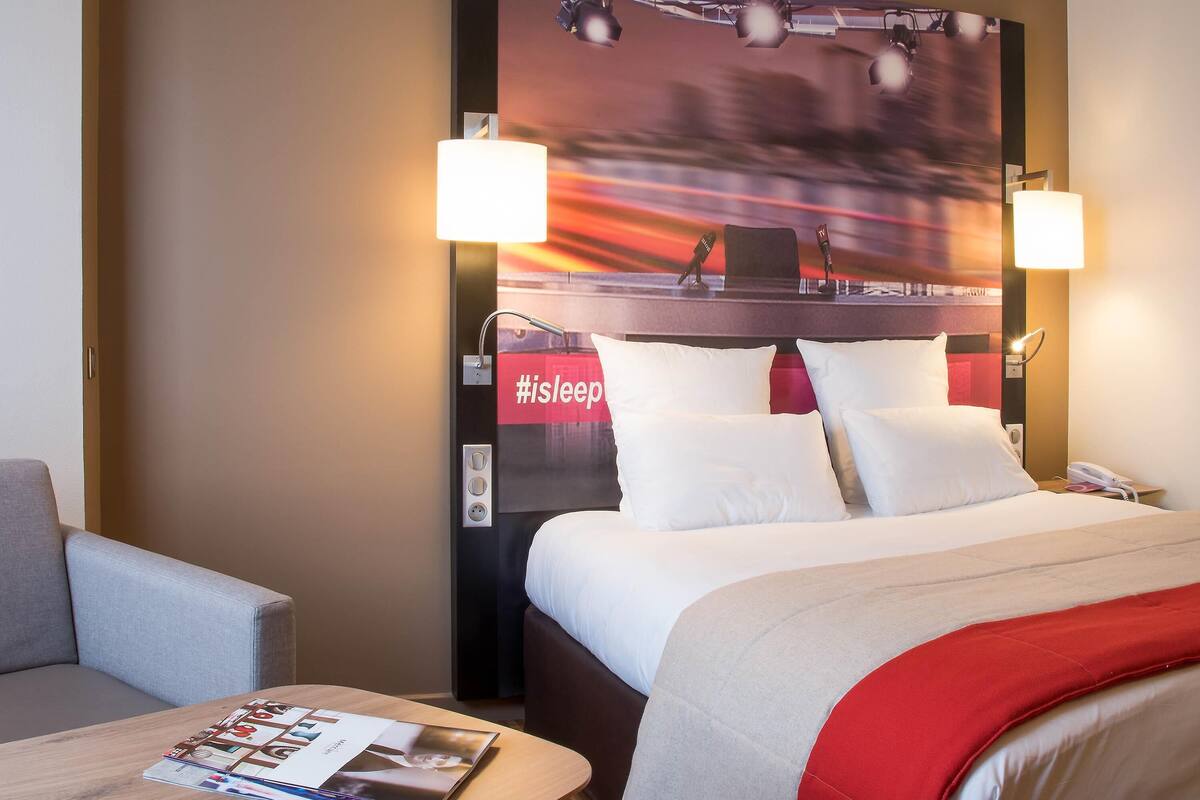 A hotel room with a bed, two bedside lamps, a nightstand with a phone, a magazine on a side table, and a large poster on the wall.