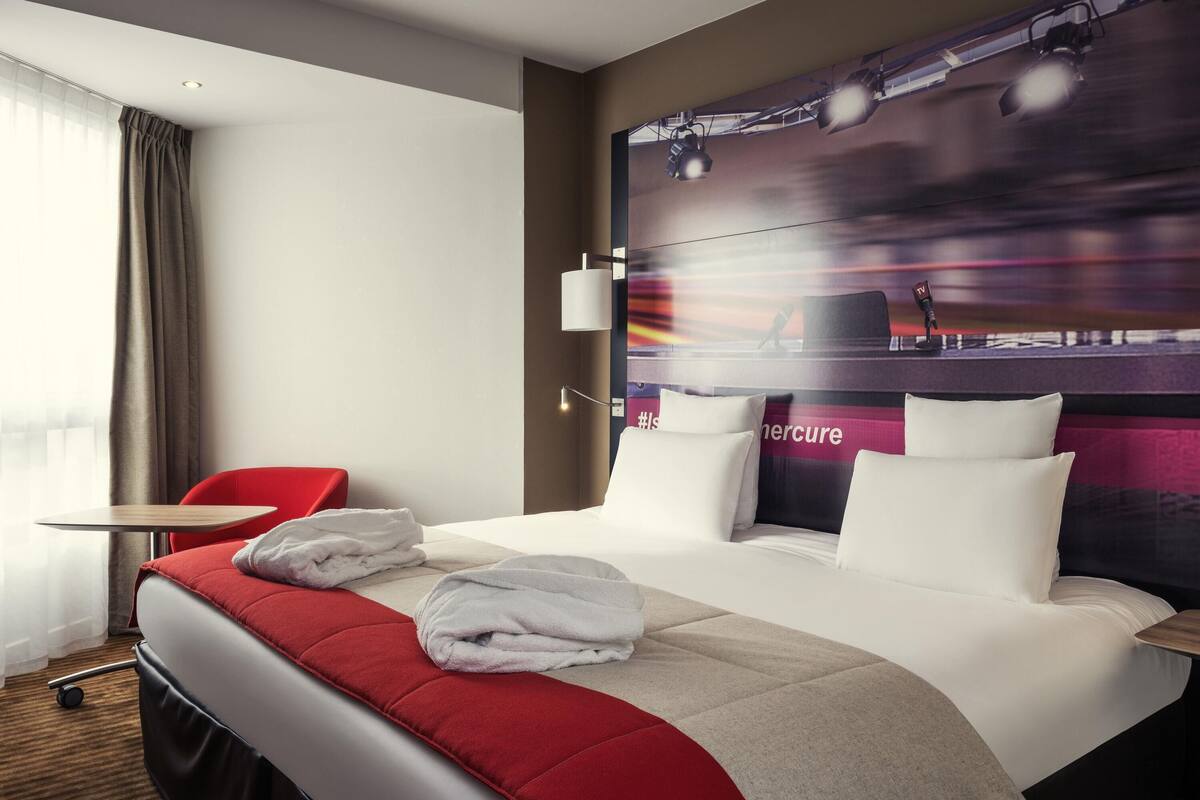 A hotel room with a large bed, a red chair, a desk, and a wall with a blurred image.