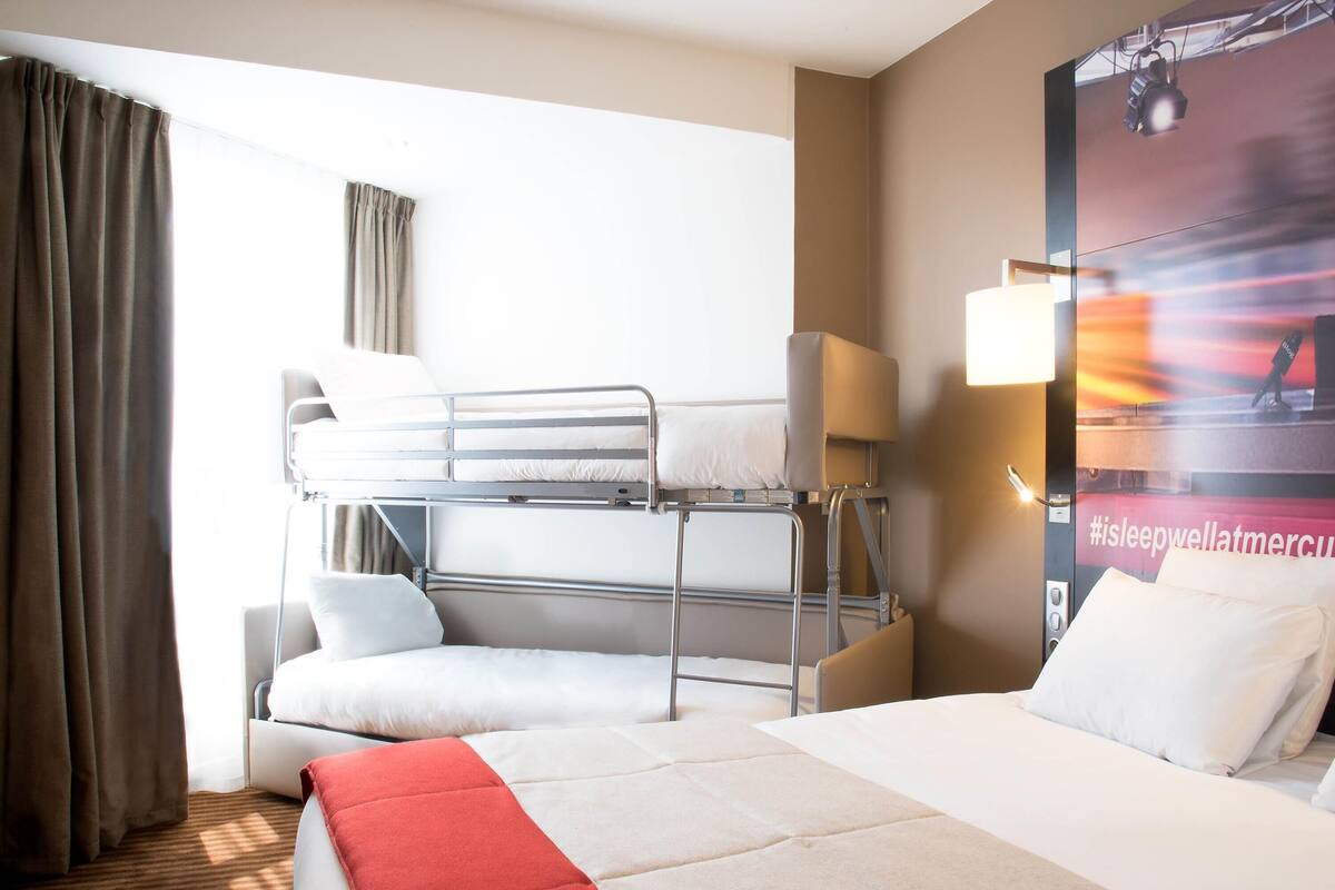 A hotel room with a bunk bed, a single bed, a red and white blanket, a lamp, and a wall with a poster.