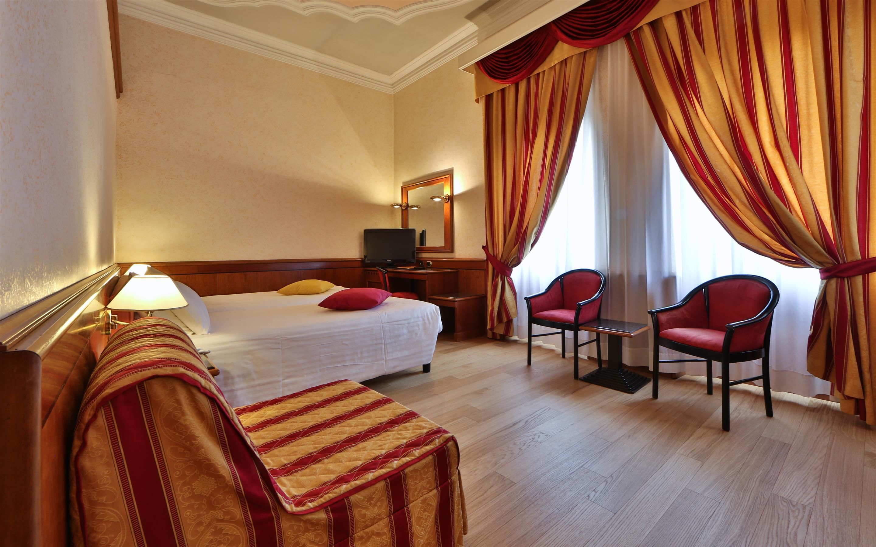 Habitación de hotel con cama, escritorio, computadora, dos sillas y ventana con cortinas.