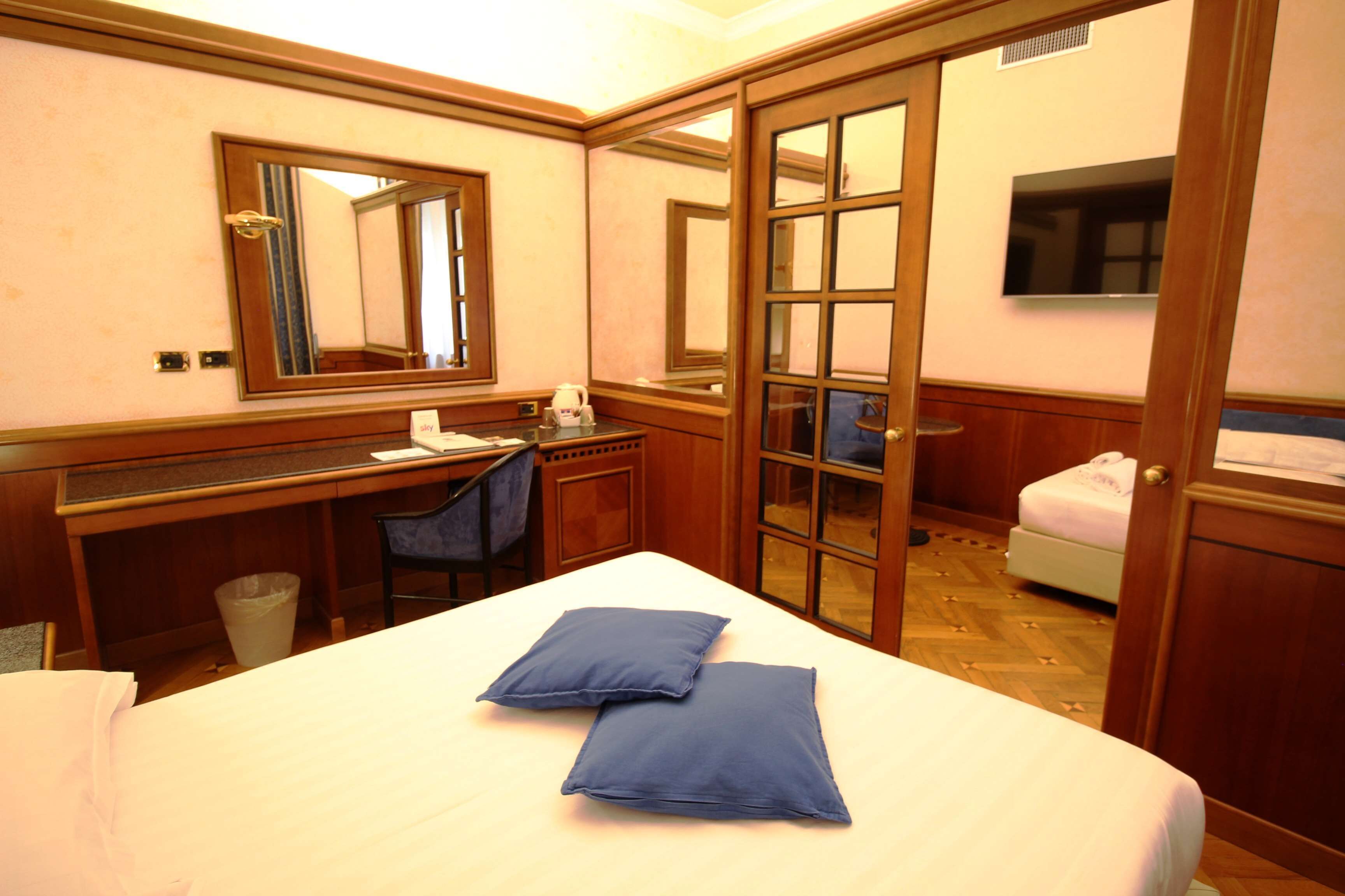 A hotel room with a bed, a desk, a chair, a small table with a kettle, a trash bin, and a TV on the wall.
