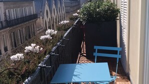Un balcón con una mesa y sillas azules, con vistas a un paisaje urbano con edificios y un árbol.