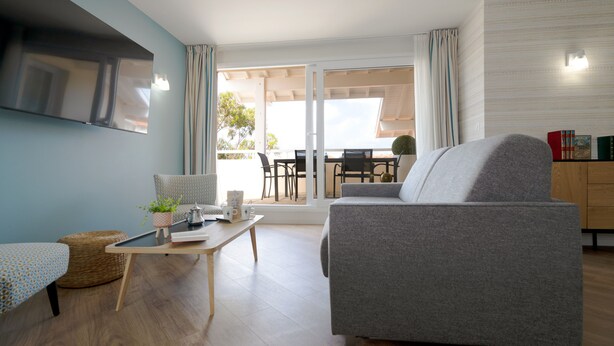 A modern living room with a grey sofa, a small table with a plant, and a TV mounted on the wall. There is a dining area with a table and chairs visible through the glass doors. - Hôtel Jules Verne Biarritz (Biarritz)