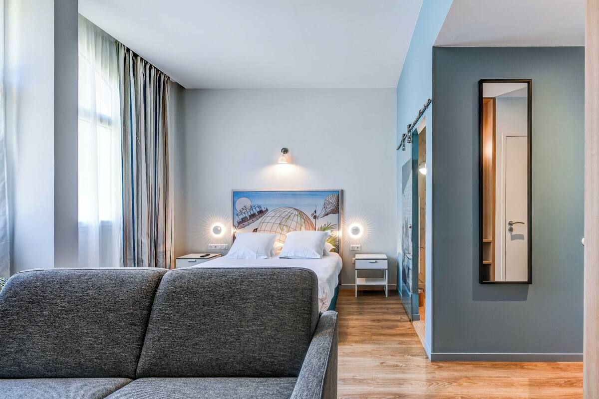 A modern hotel room with a large bed, a sofa, a painting on the wall, and a mirror.