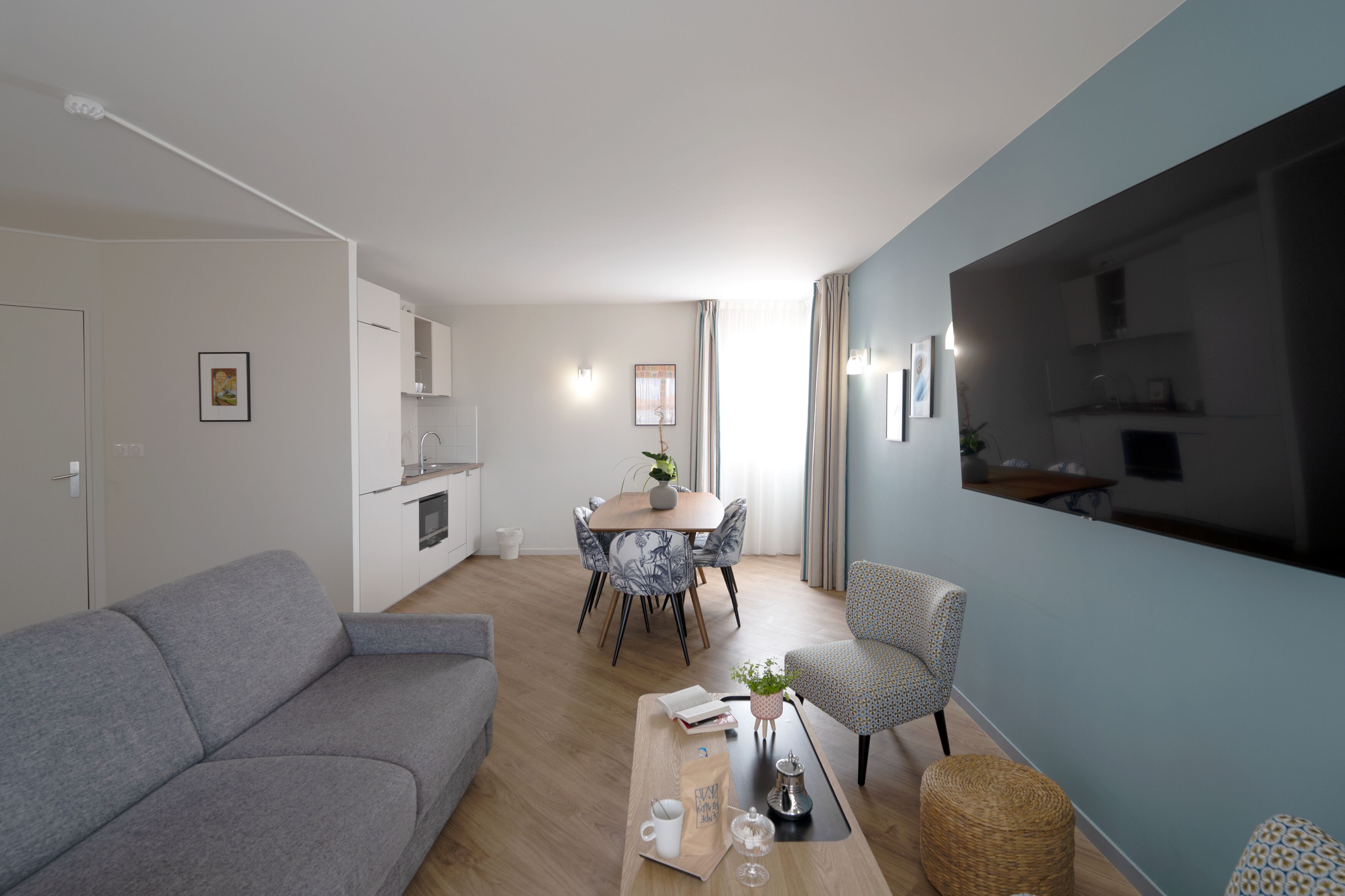 En moderne stue med en grå sofa, en mønstret lænestol, et spisebord med stole, og et køkken i baggrunden.